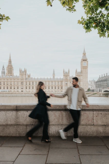 London couple photoshoot walking beneath riverside trees Couple walking hand in hand beside the Thames beneath green tree branches, with Parliament behind them, during their London photoshoot.