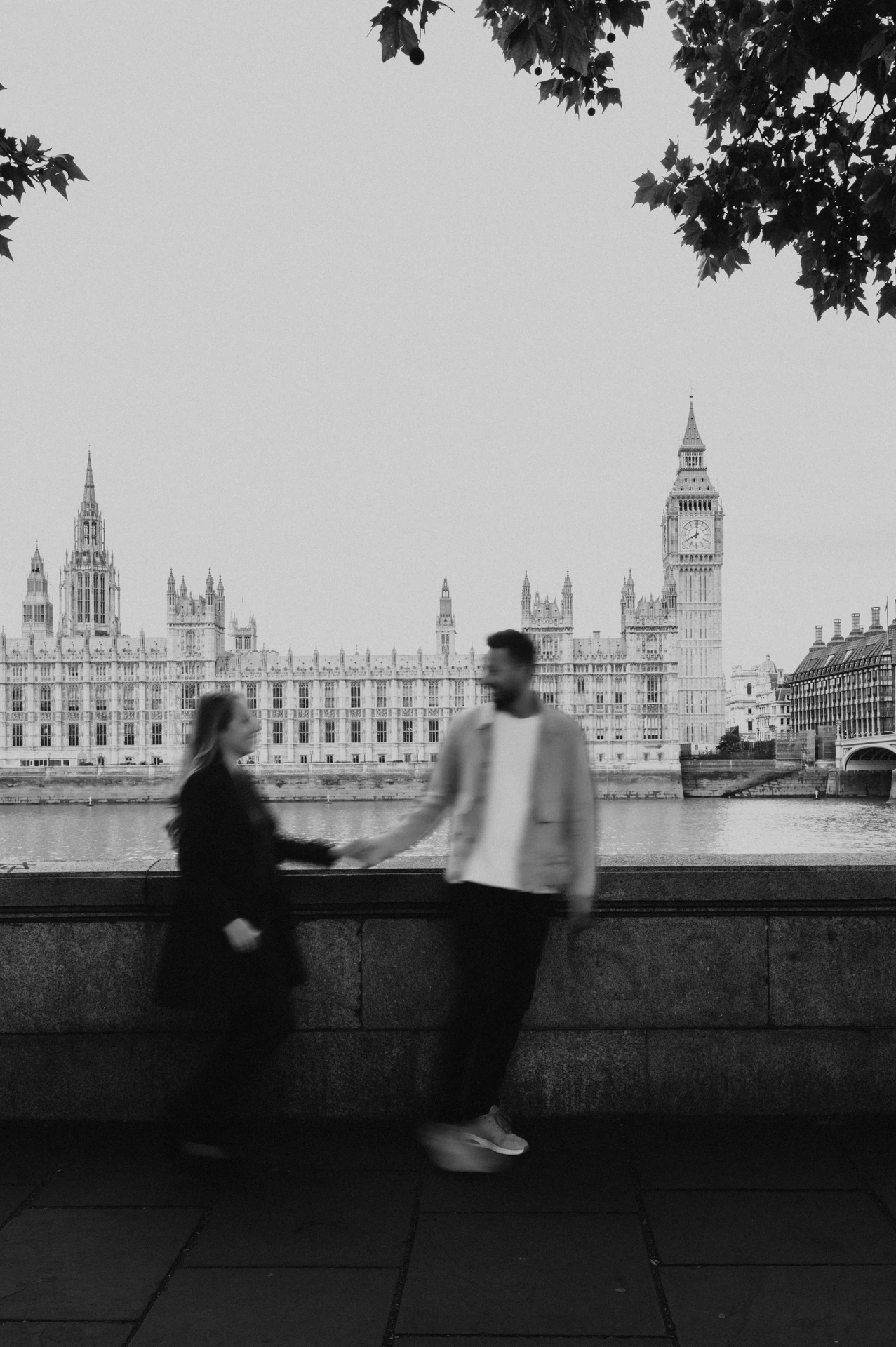 Black-and-white photograph of a couple walking hand in hand beneath tree branches beside the Thames during their London photoshoot.