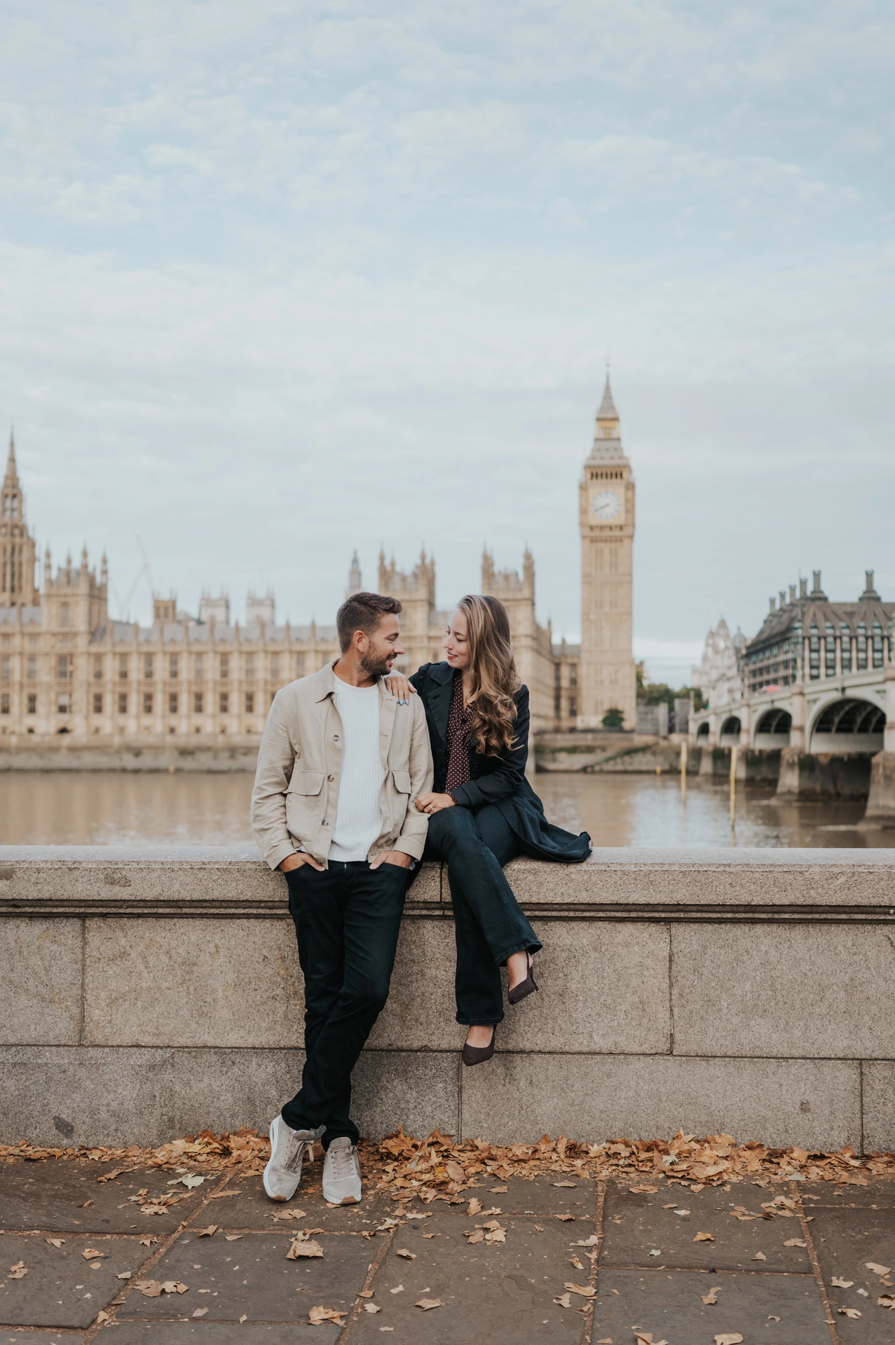Couple smiling at one another beside the stone riverside wall, with Big Ben across the Thames during their London couple photoshoot.