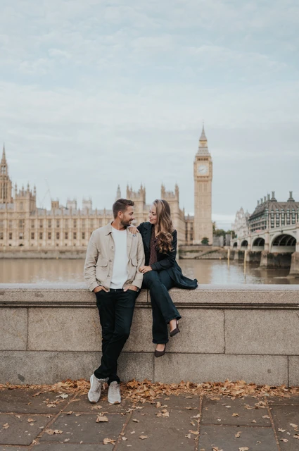 Couple smiling at one another beside the stone riverside wall, with Big Ben across the Thames during their London couple photoshoot.