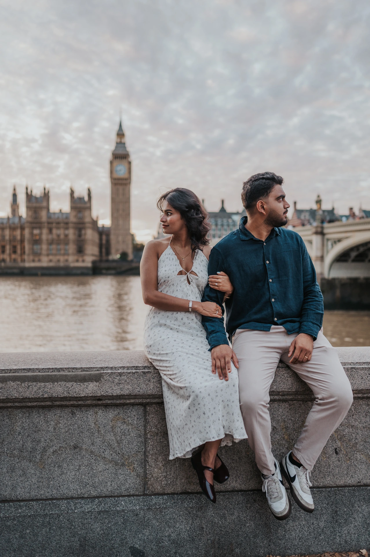 Couple seated together on the stone riverside wall and looking in opposite directions, with Big Ben behind them, during their London couple photoshoot.
