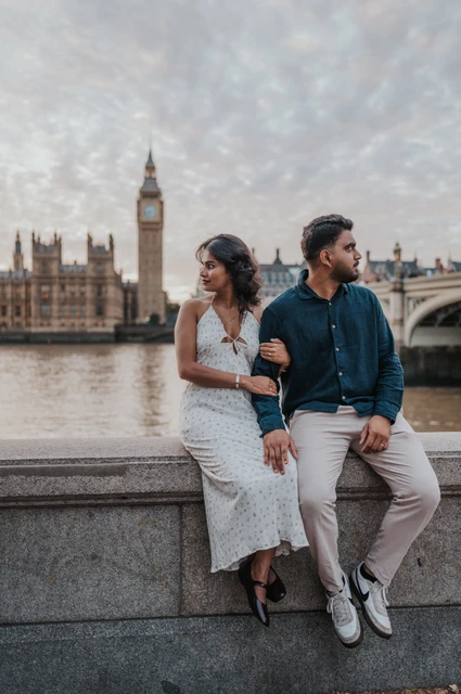 Couple seated together on the stone riverside wall and looking in opposite directions, with Big Ben behind them, during their London couple photoshoot.