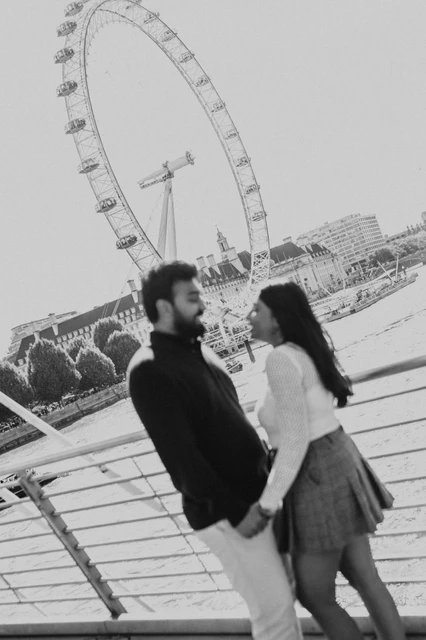 An angled black-and-white bridge portrait — London couple photoshoot An angled black-and-white portrait of the couple holding hands and facing each other on Golden Jubilee Bridge, with the London Eye beyond the railing during their London couple photoshoot.