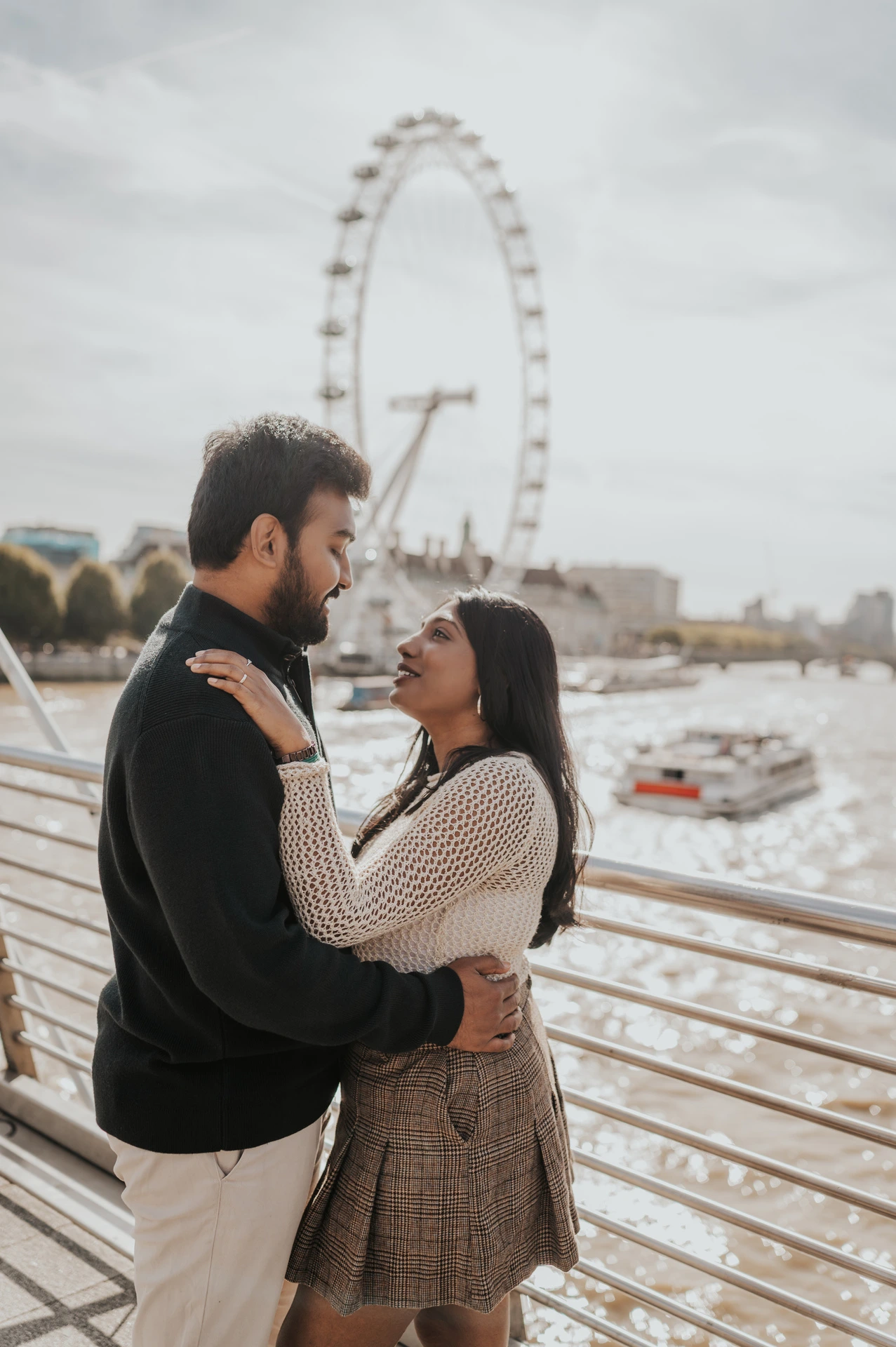 The couple embracing beside the Golden Jubilee Bridge railing, with her hands at his shoulders and the London Eye behind them during their London couple photoshoot.