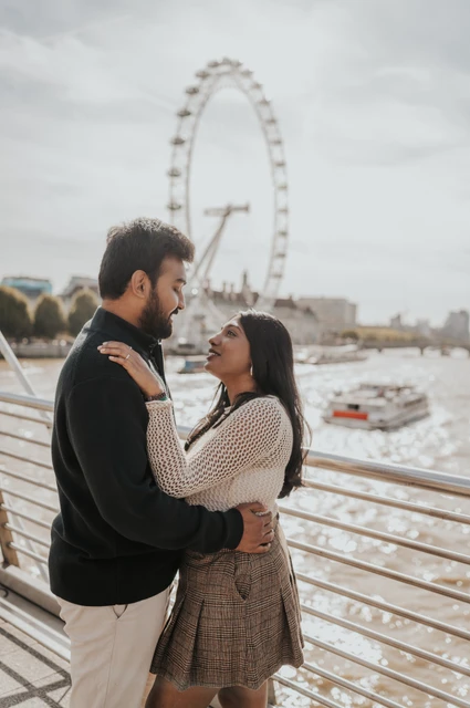 An embrace above the Thames — London couple photoshoot The couple embracing beside the Golden Jubilee Bridge railing, with her hands at his shoulders and the London Eye behind them during their London couple photoshoot.