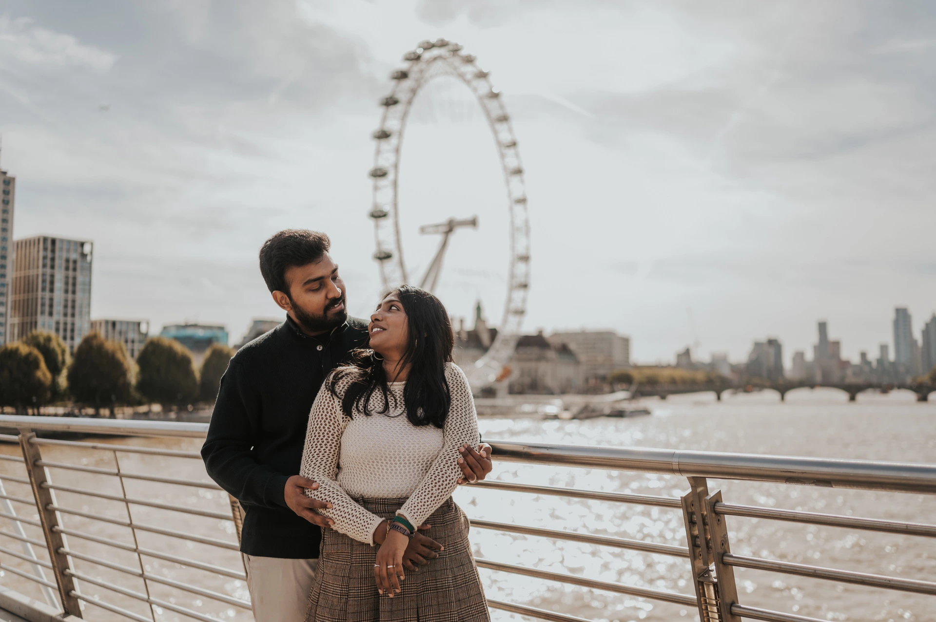 The couple standing together at the Golden Jubilee Bridge railing, with the man behind the woman and the London Eye across the river during their London couple photoshoot.