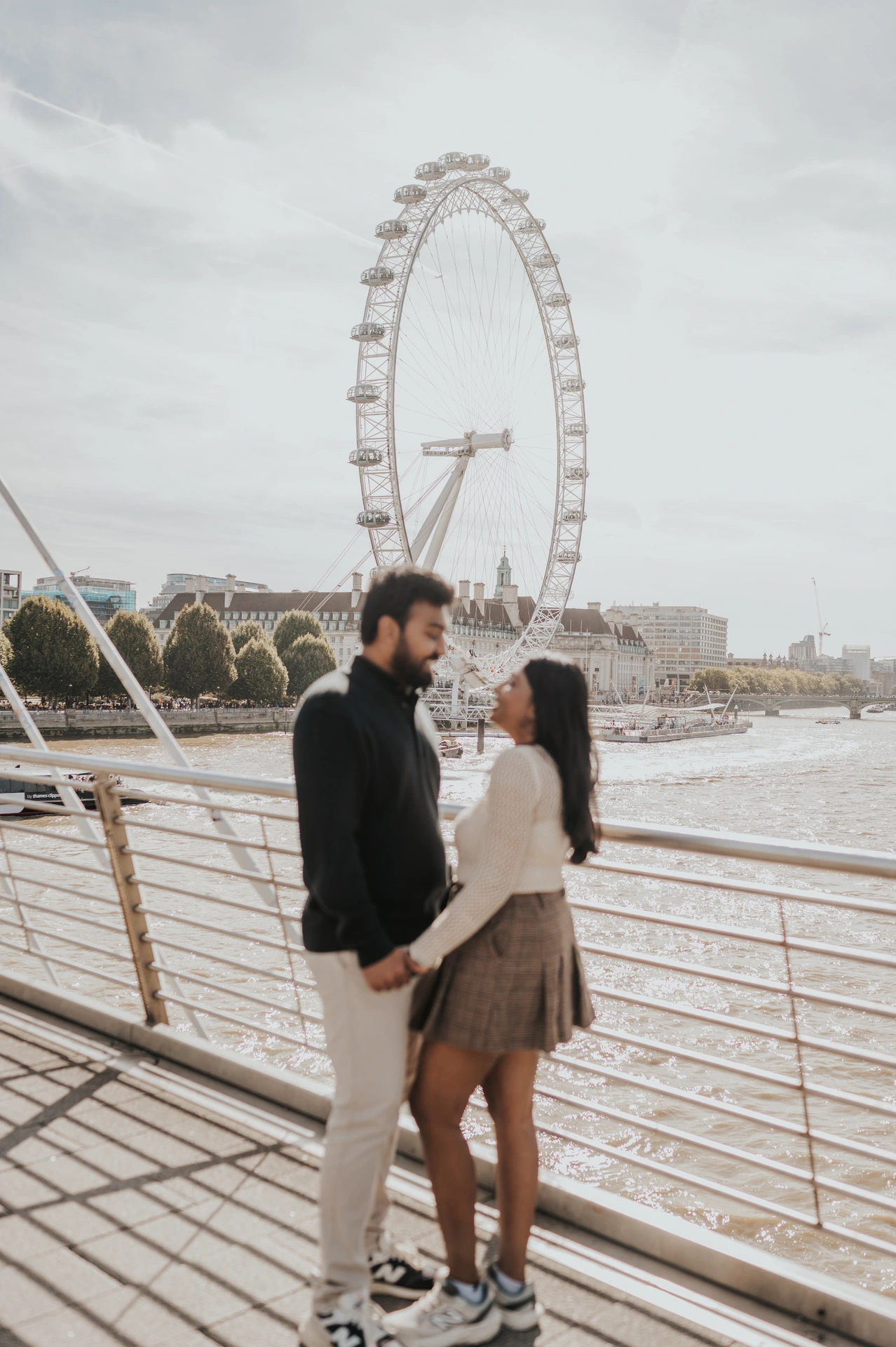 The couple facing one another and holding hands beside the Golden Jubilee Bridge railing, framed beneath the London Eye during their London couple photoshoot.