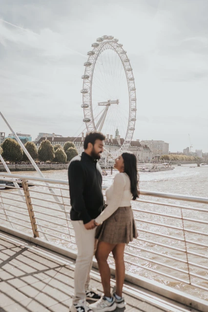 Facing each other on Golden Jubilee Bridge — London couple photoshoot The couple facing one another and holding hands beside the Golden Jubilee Bridge railing, framed beneath the London Eye during their London couple photoshoot.