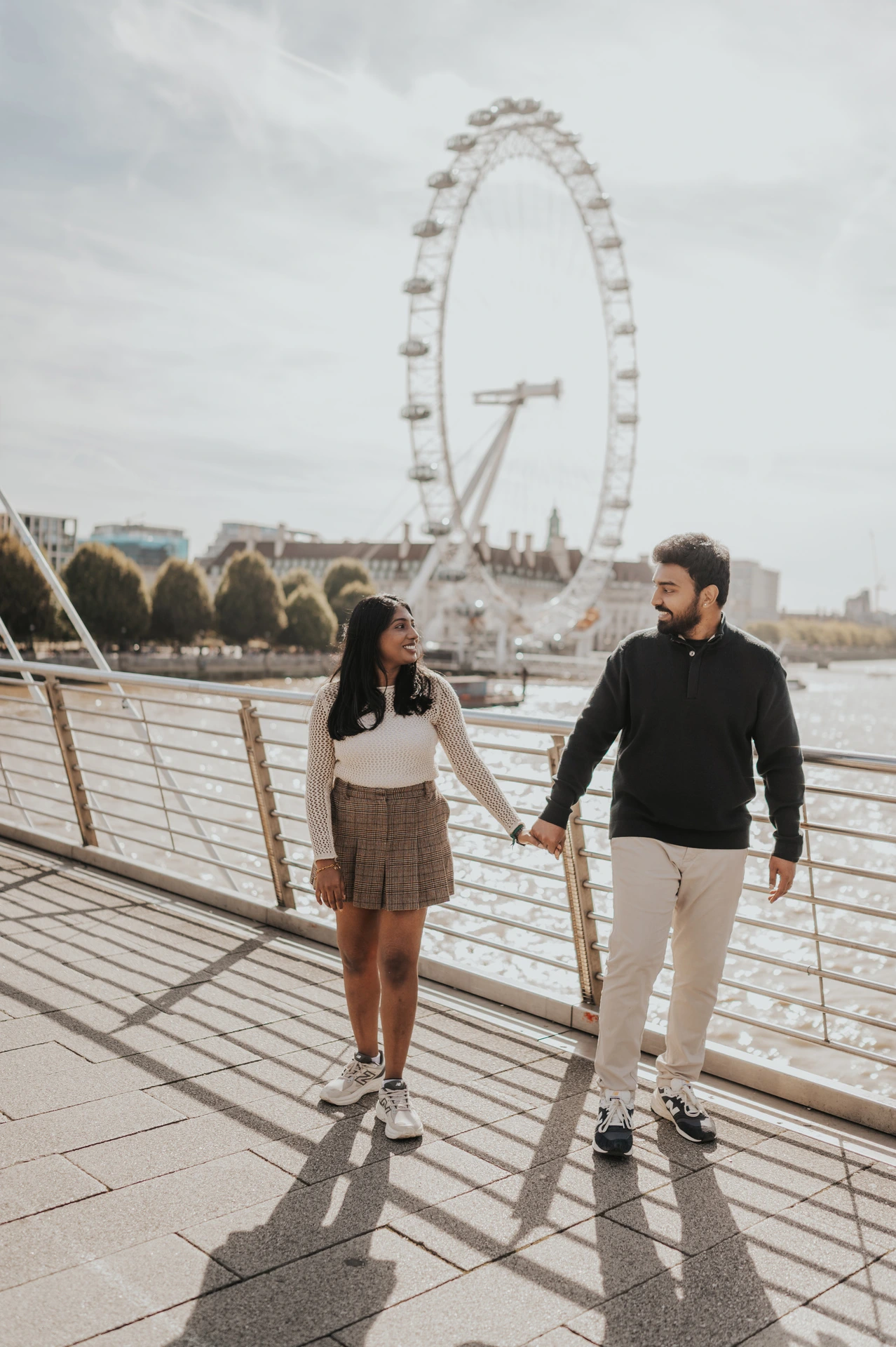 The couple holding hands on Golden Jubilee Bridge, with their full-length shadows on the walkway and the London Eye across the Thames during their London couple photoshoot.