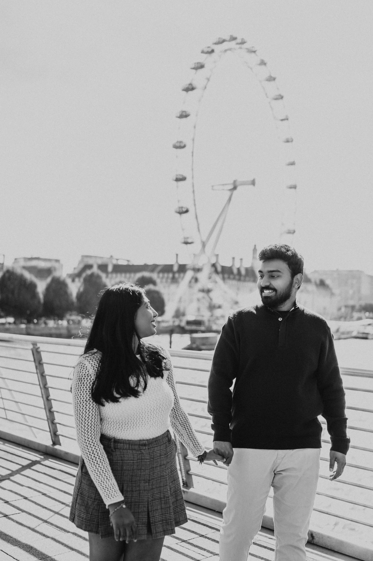 A black-and-white portrait of the couple holding hands and looking at one another on Golden Jubilee Bridge beneath the London Eye during their London couple photoshoot.