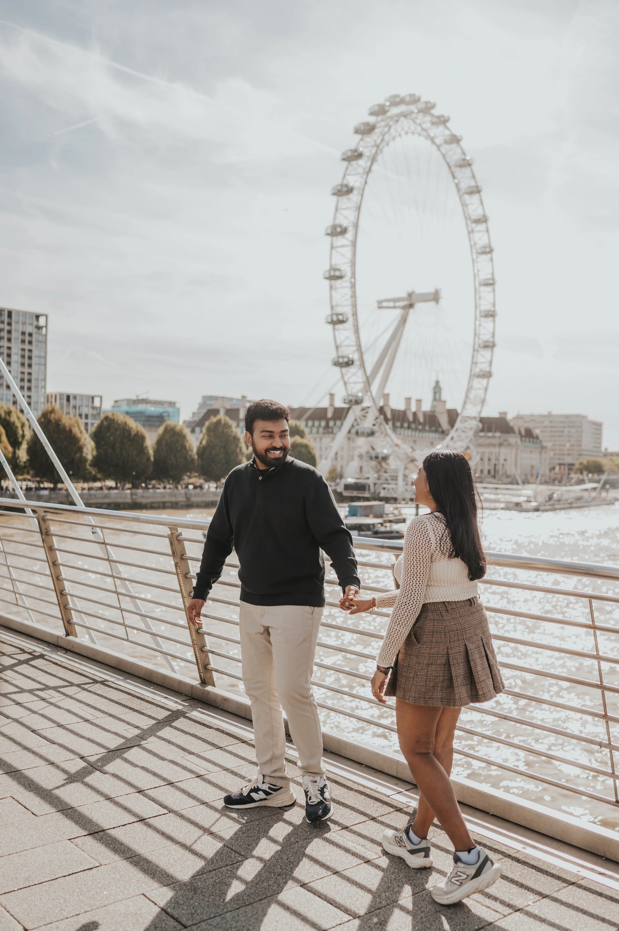 A full-length portrait of the couple holding hands on Golden Jubilee Bridge, with the woman turned towards the man and the London Eye behind them during their London couple photoshoot.