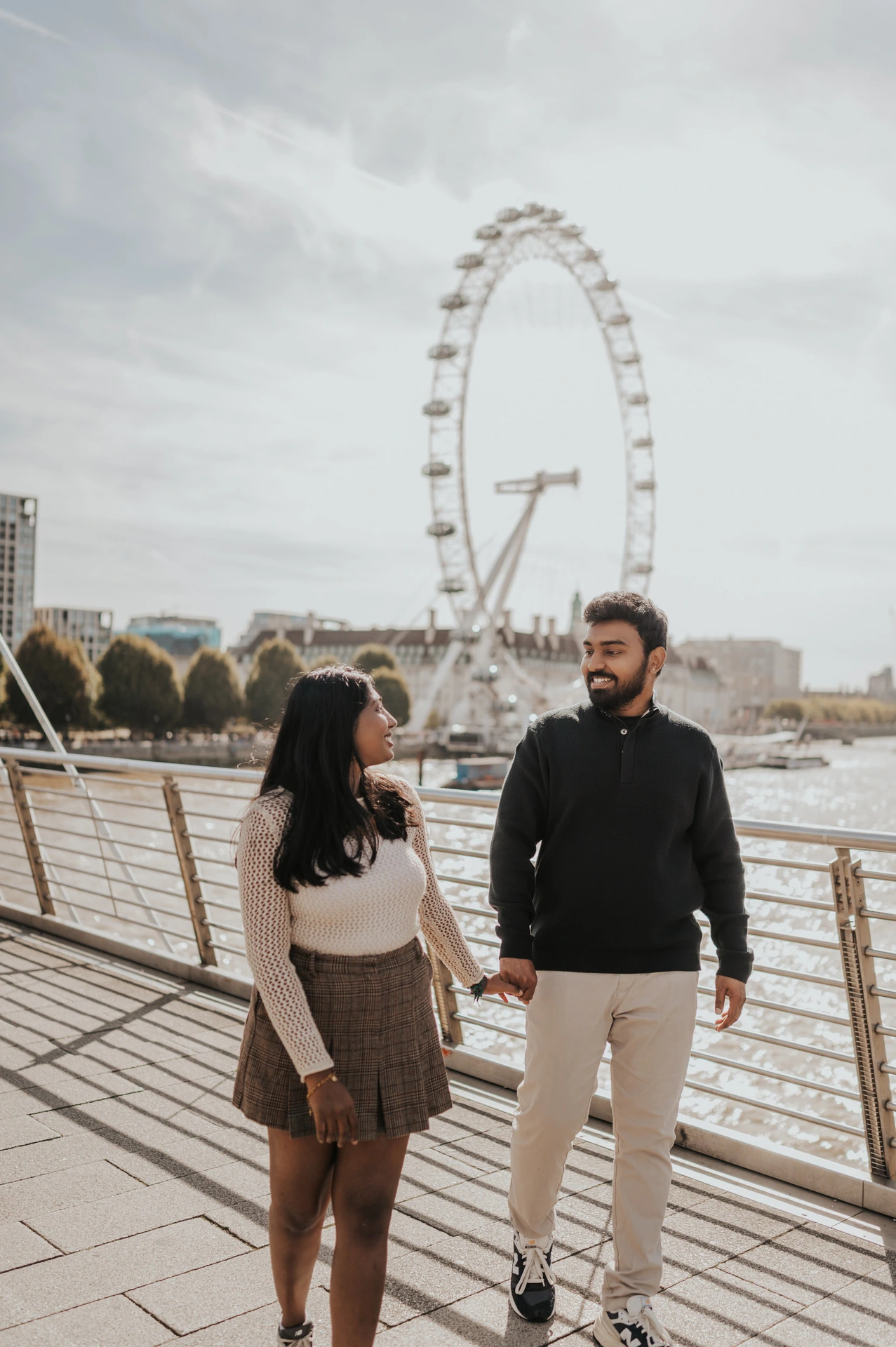 A full-length view of the couple holding hands and looking at each other on Golden Jubilee Bridge, with the London Eye behind them during their London couple photoshoot.
