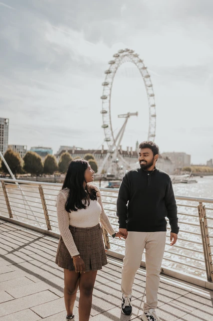 Holding hands on Golden Jubilee Bridge — London couple photoshoot A full-length view of the couple holding hands and looking at each other on Golden Jubilee Bridge, with the London Eye behind them during their London couple photoshoot.