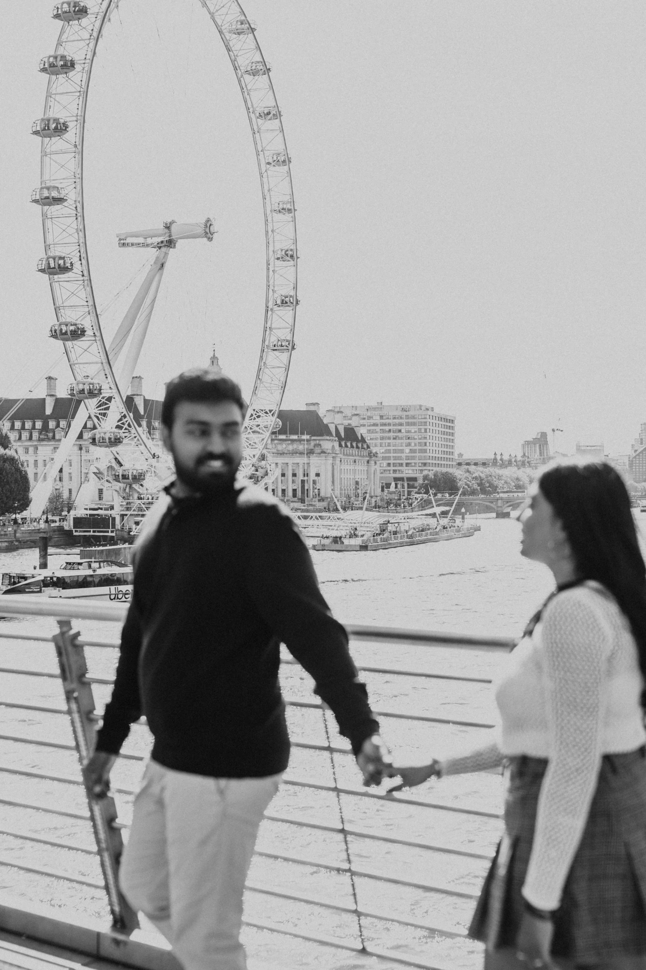 A black-and-white portrait of the man looking towards the camera while holding the woman’s hand on Golden Jubilee Bridge, with the London Eye behind them during their London couple photoshoot.