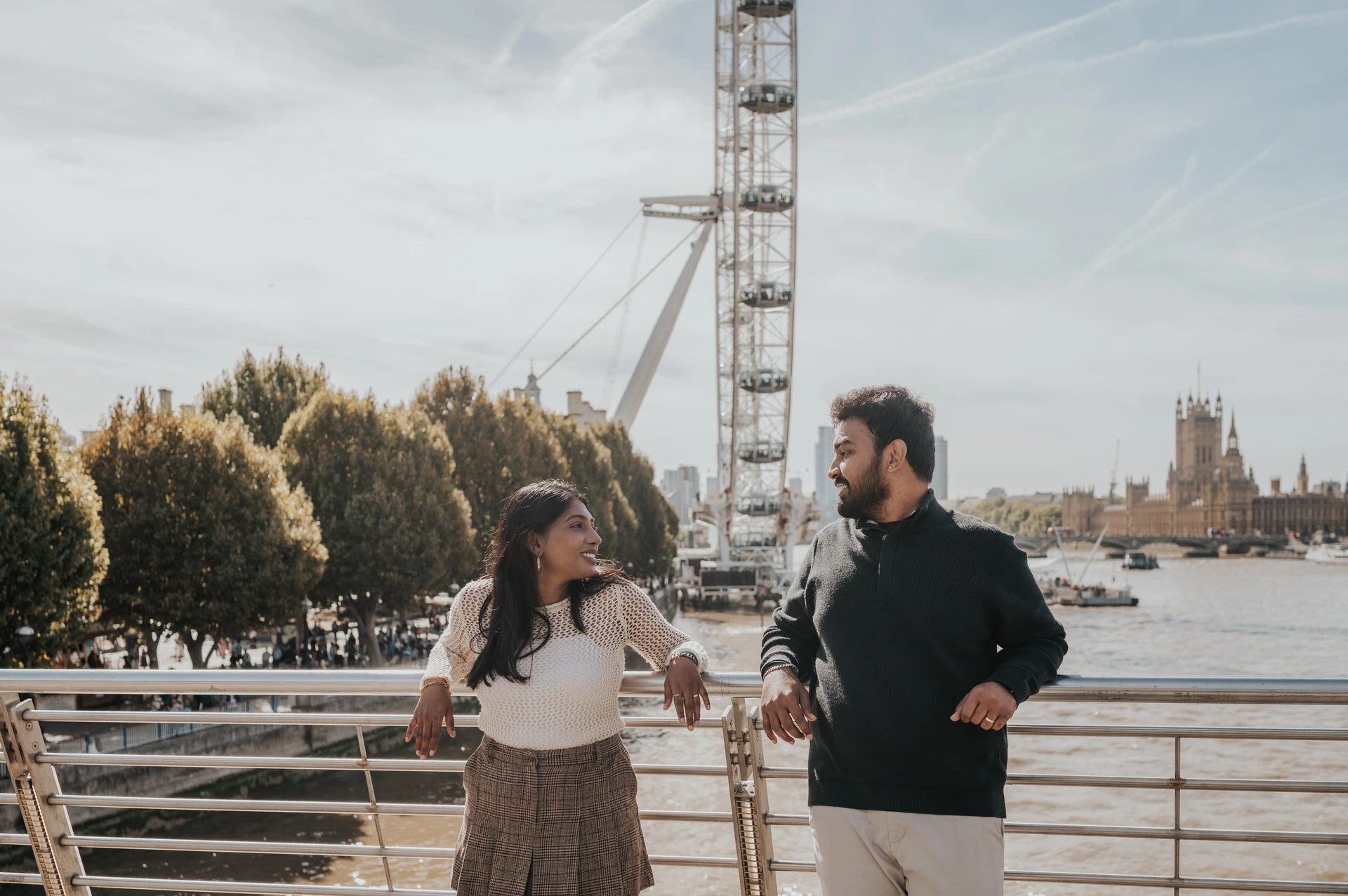 A wide portrait of the couple looking at each other by the Golden Jubilee Bridge railing, with the London Eye, trees and Thames behind them during their London couple photoshoot.