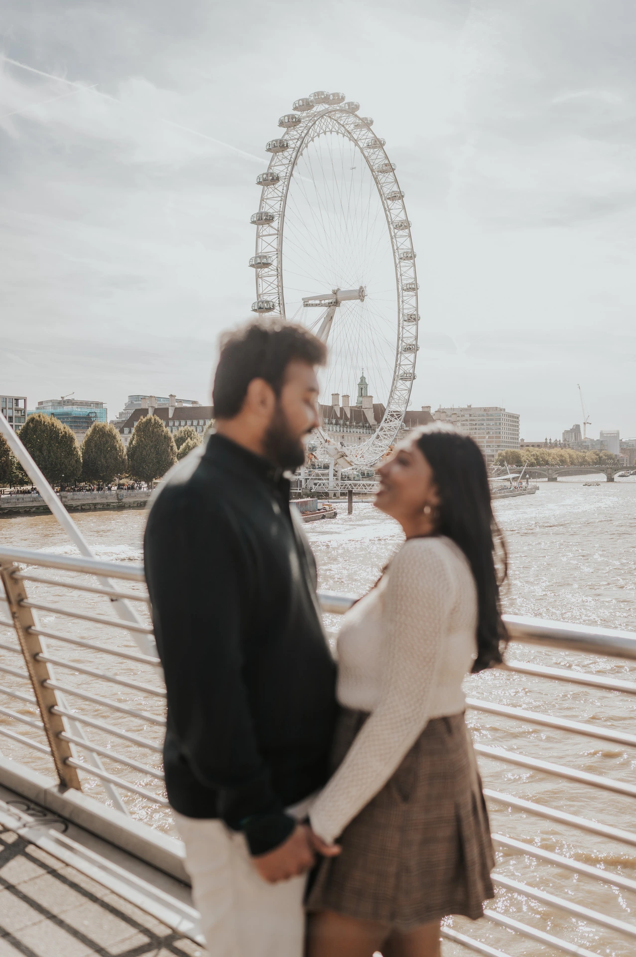 A shallow-focus portrait of the couple facing each other on Golden Jubilee Bridge, with the man in the foreground and the London Eye across the water during their London couple photoshoot.