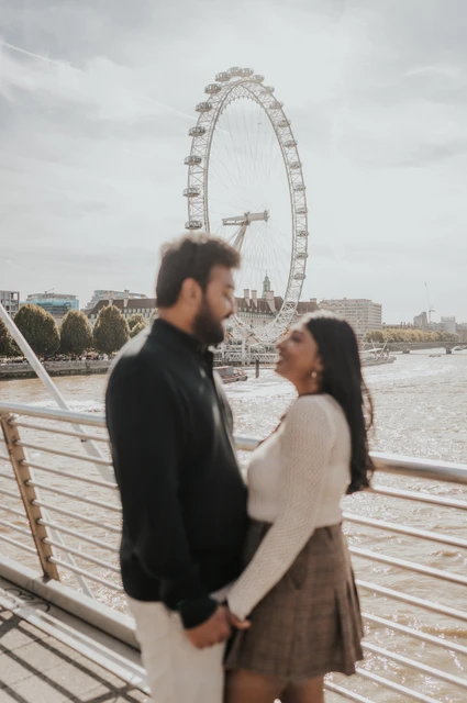 A shallow-focus portrait by the bridge railing — London couple photoshoot A shallow-focus portrait of the couple facing each other on Golden Jubilee Bridge, with the man in the foreground and the London Eye across the water during their London couple photoshoot.