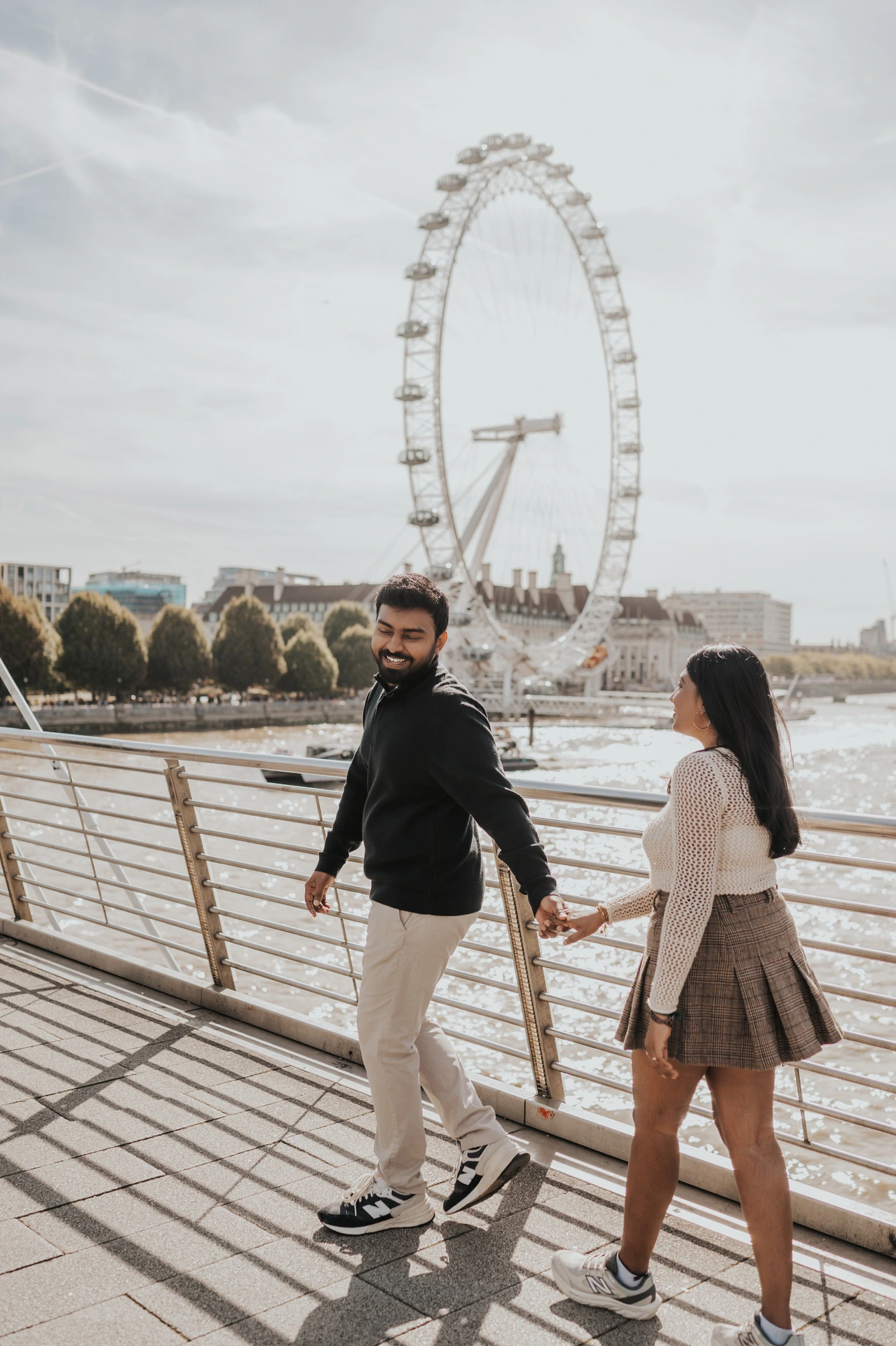 The couple holding hands as the man looks back on Golden Jubilee Bridge, with the London Eye above the opposite riverbank during their London couple photoshoot.