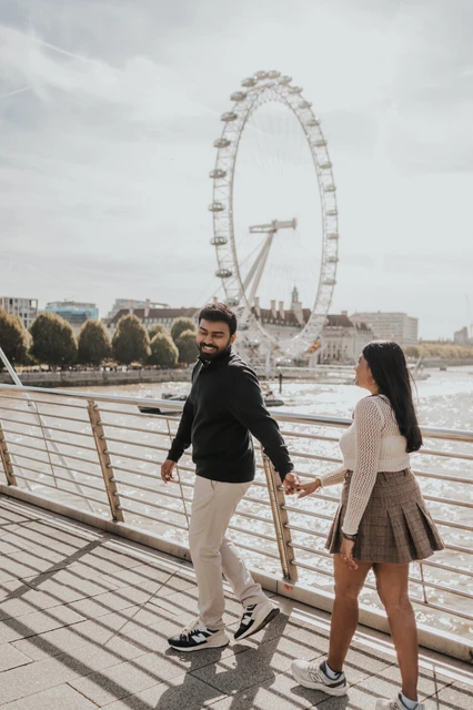 A look back beside the London Eye — London couple photoshoot The couple holding hands as the man looks back on Golden Jubilee Bridge, with the London Eye above the opposite riverbank during their London couple photoshoot.