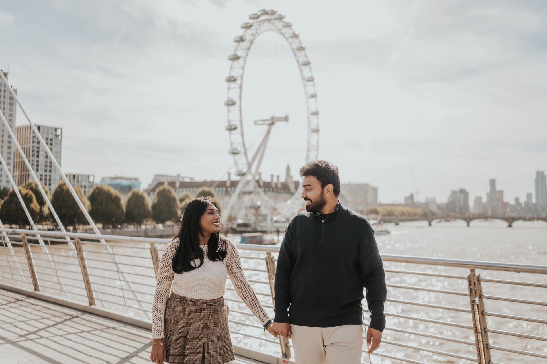 A wide view across the Thames from the bridge — London couple photoshoot A wide view of the couple holding hands beside the Golden Jubilee Bridge railing, with the London Eye and the river behind them during their London couple photoshoot.