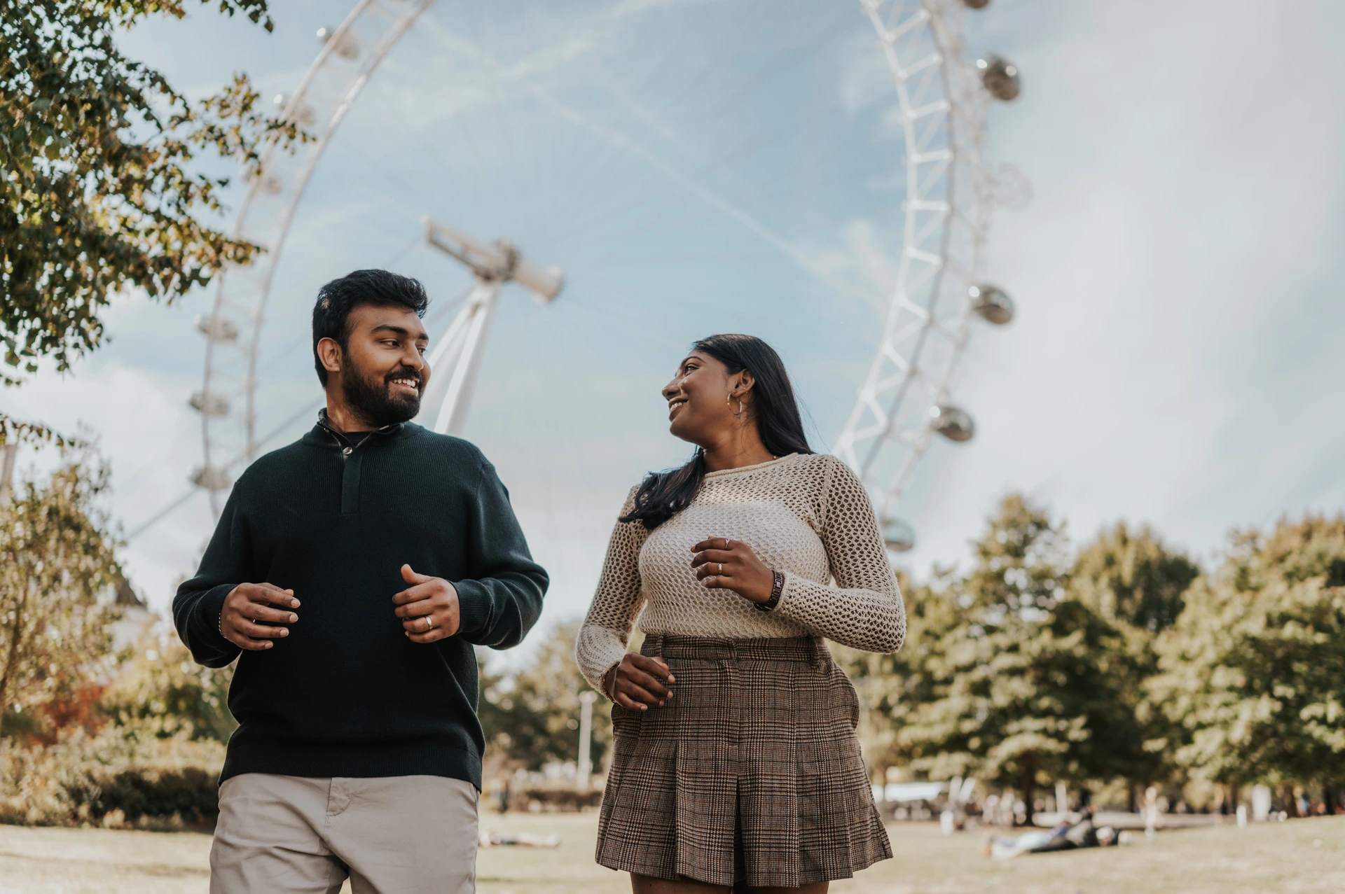 The couple looking at one another in a closer landscape portrait on the Jubilee Gardens lawn, with the London Eye and leafy trees behind them during their London couple photoshoot.