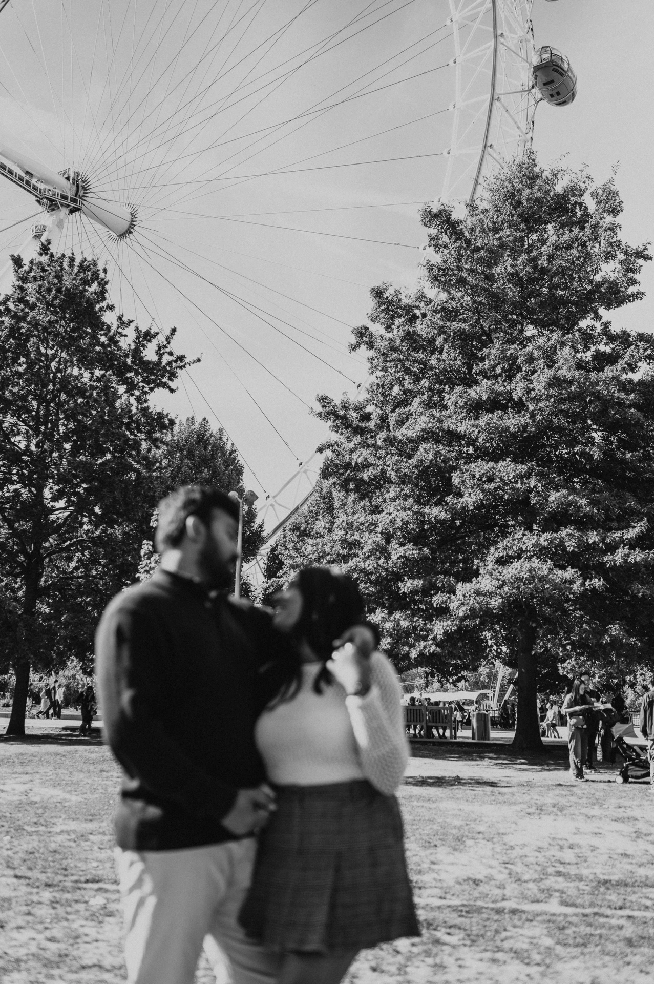 A black-and-white full-length portrait of the couple embracing on the Jubilee Gardens lawn, with trees and part of the London Eye overhead during their London couple photoshoot.