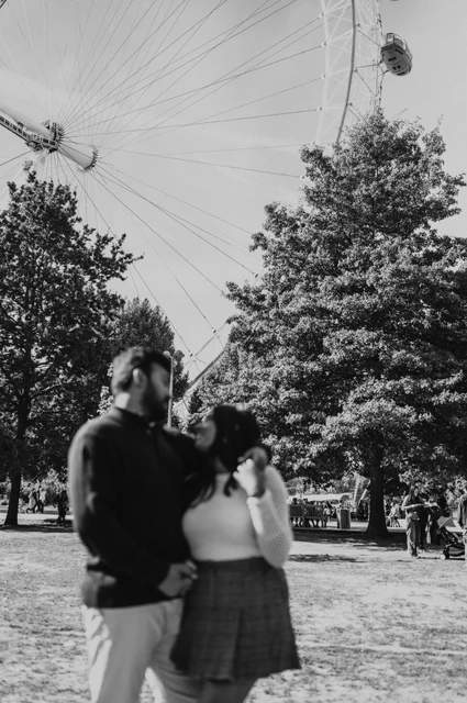 An embrace beneath the wheel in Jubilee Gardens — London couple photoshoot A black-and-white full-length portrait of the couple embracing on the Jubilee Gardens lawn, with trees and part of the London Eye overhead during their London couple photoshoot.