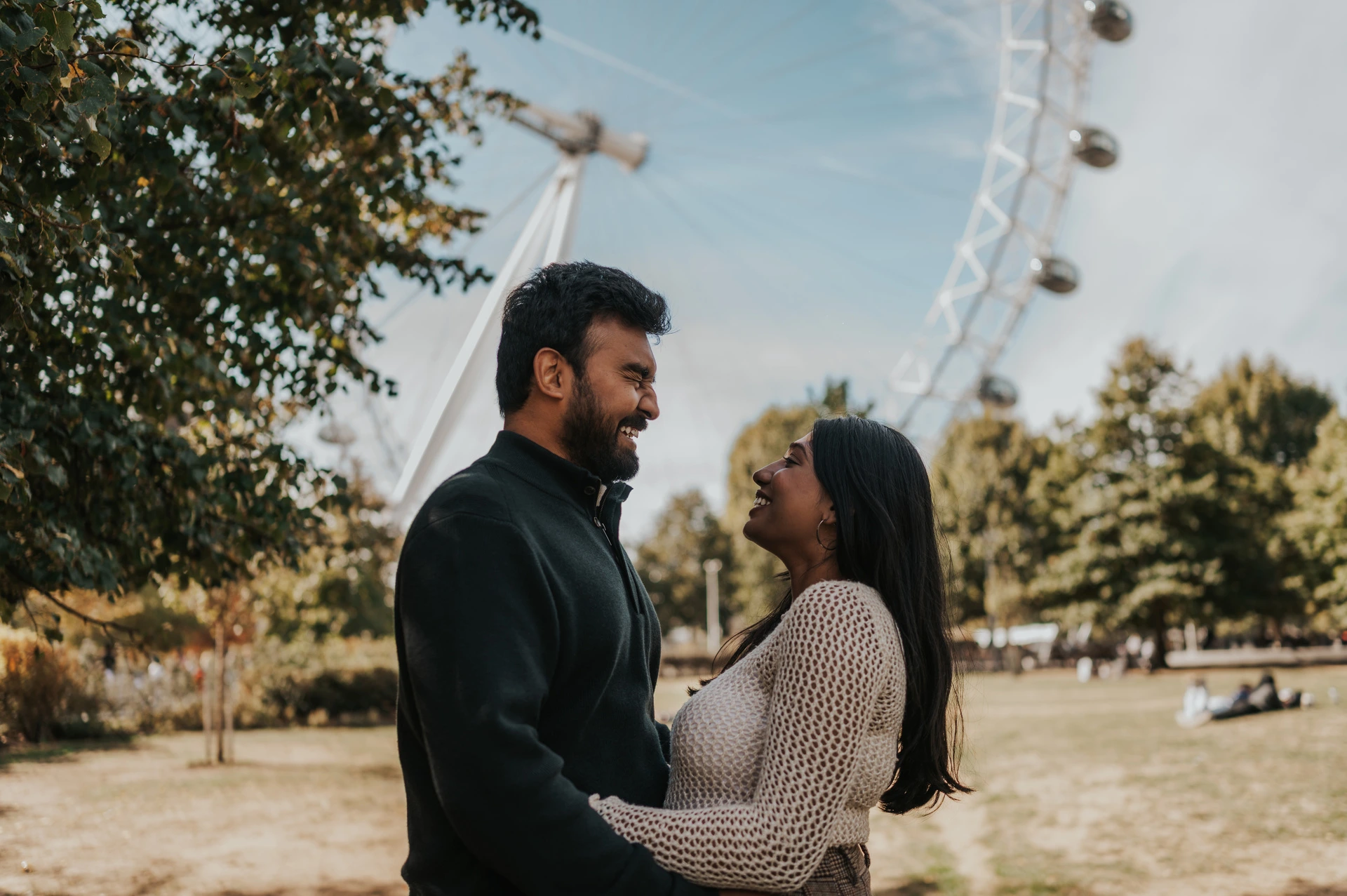 The couple facing each other and holding one another at the waist on the Jubilee Gardens lawn, with leafy trees and the London Eye behind them during their London couple photoshoot.