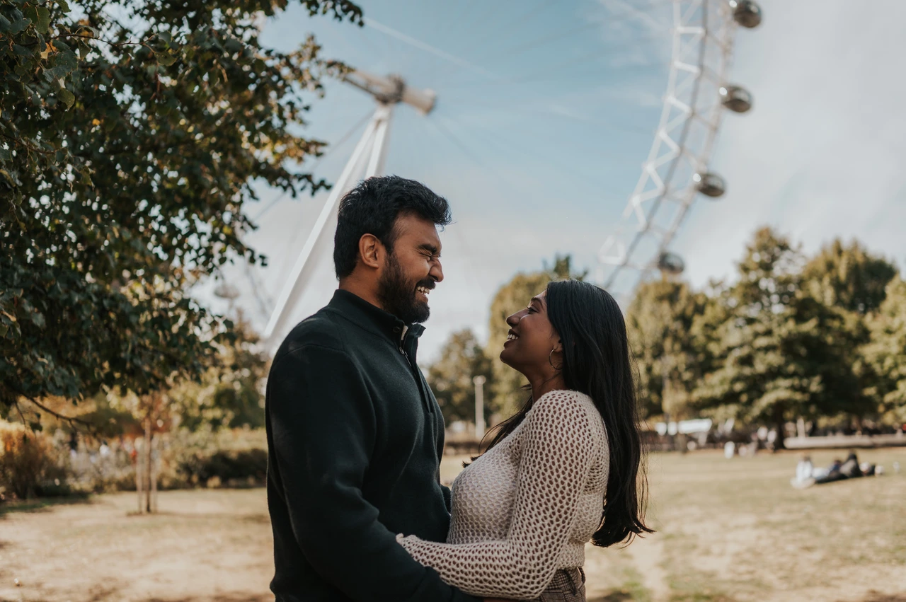 Facing each other on the Jubilee Gardens lawn — London couple photoshoot The couple facing each other and holding one another at the waist on the Jubilee Gardens lawn, with leafy trees and the London Eye behind them during their London couple photoshoot.