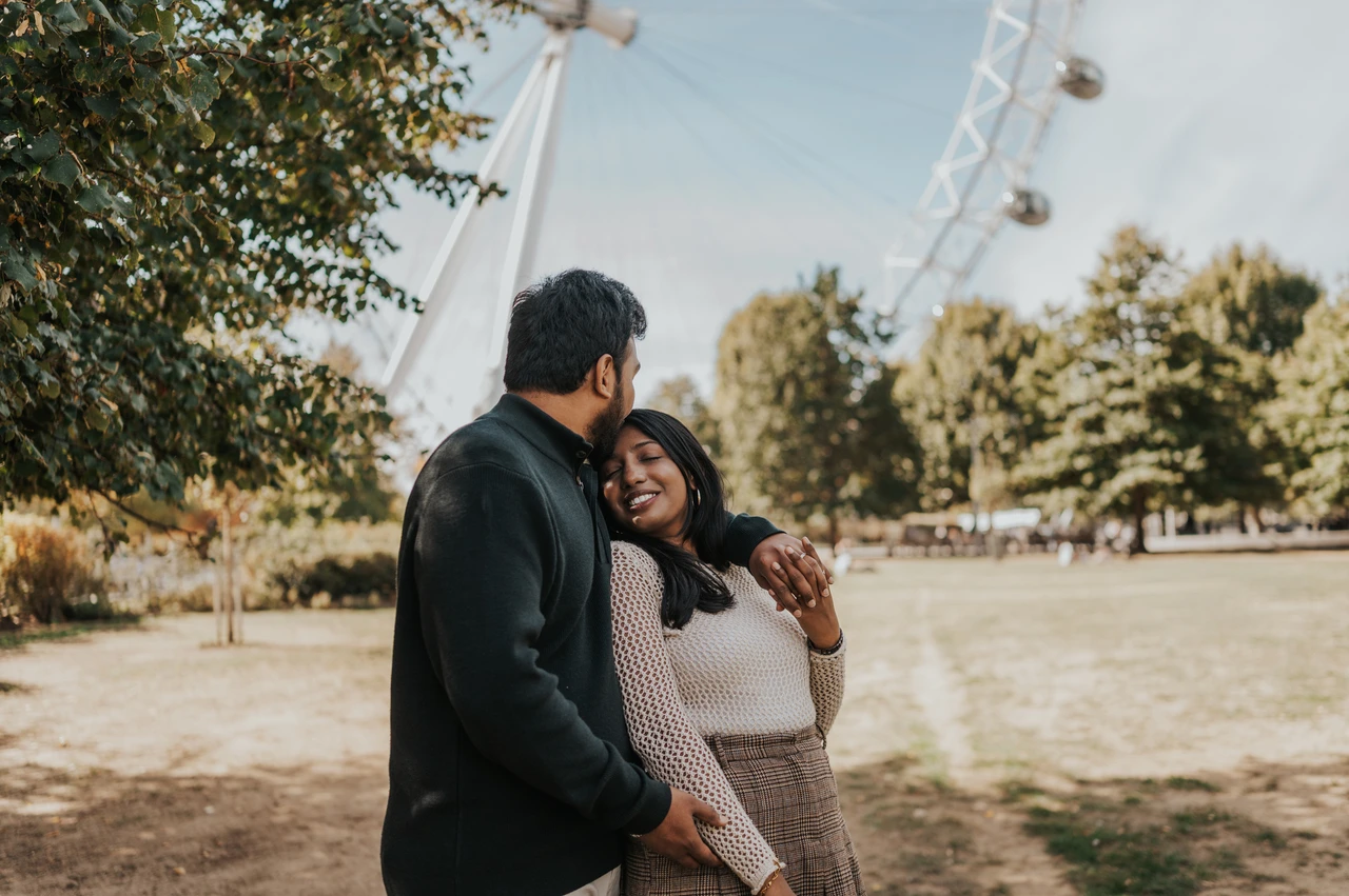 A forehead kiss in Jubilee Gardens — London couple photoshoot The man kissing the woman’s forehead as they stand together in Jubilee Gardens, with trees and part of the London Eye behind them during their London couple photoshoot.