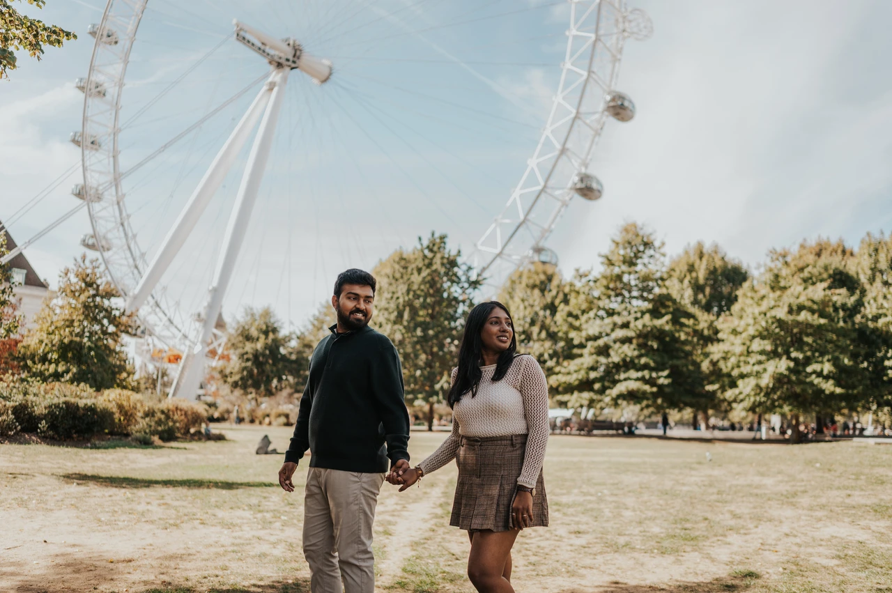 Holding hands under the London Eye in Jubilee Gardens — London couple photoshoot The couple holding hands on the Jubilee Gardens lawn, with the London Eye curving above them and trees in the background during their London couple photoshoot.