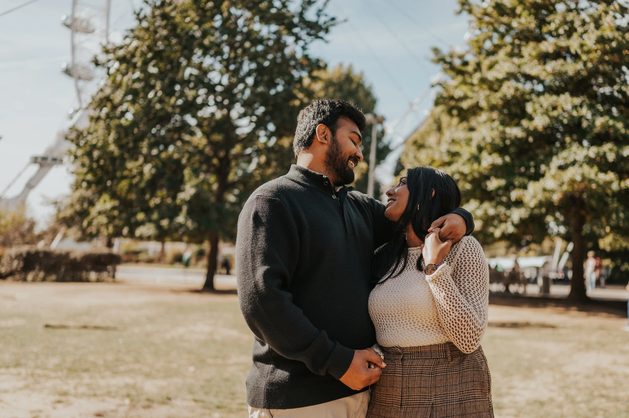 A smiling portrait among the trees in Jubilee Gardens — London couple photoshoot The couple smiling at one another in Jubilee Gardens, with his arm around her shoulders and leafy trees behind them during their London couple photoshoot.