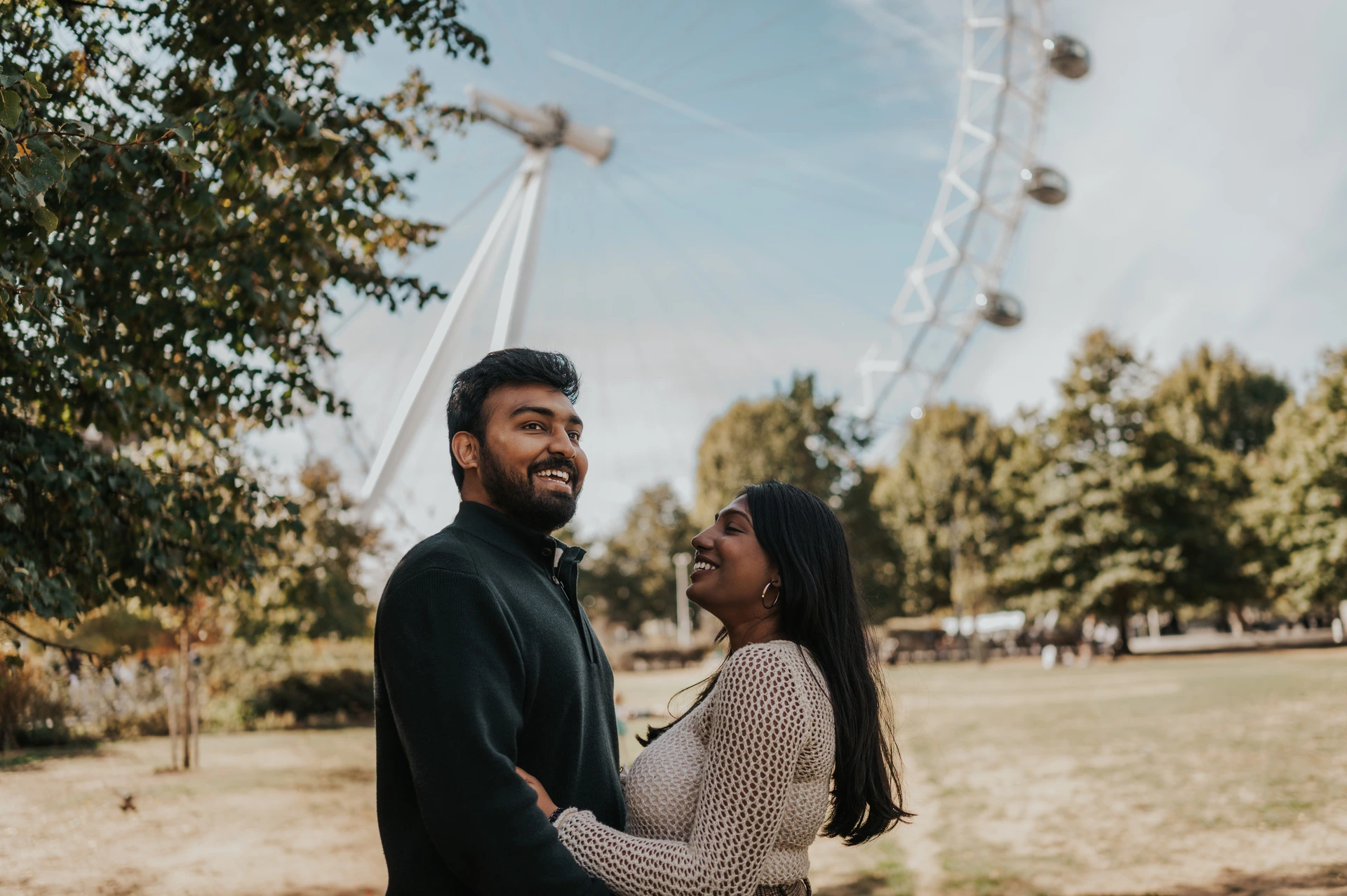 Smiles beneath the London Eye in Jubilee Gardens — London couple photoshoot The couple smiling at each other beneath a leafy tree in Jubilee Gardens, with the London Eye behind them during their London couple photoshoot.