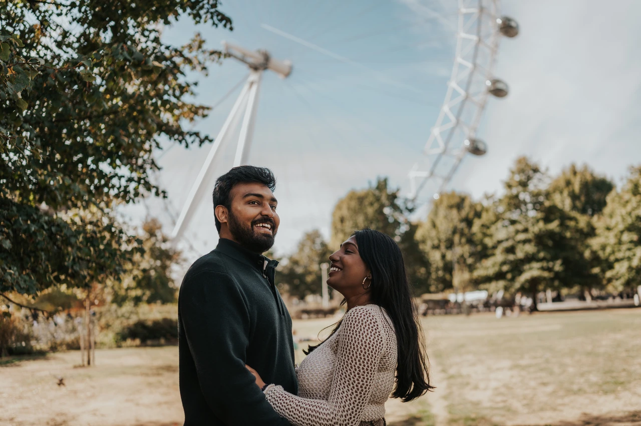 Smiles beneath the London Eye in Jubilee Gardens — London couple photoshoot The couple smiling at each other beneath a leafy tree in Jubilee Gardens, with the London Eye behind them during their London couple photoshoot.