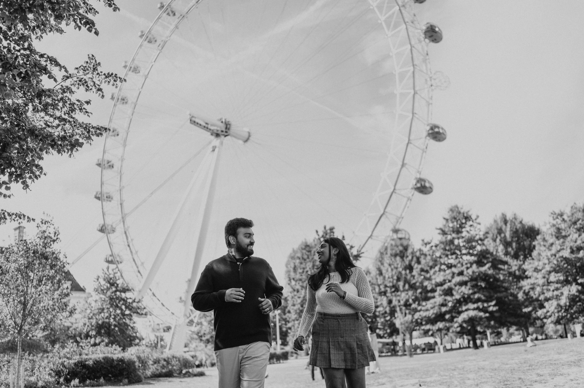 A wide black-and-white view of the couple beside one another on the Jubilee Gardens lawn, with the London Eye above the trees during their London couple photoshoot.