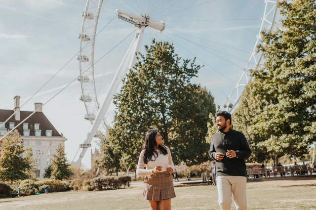 A wide portrait of the couple looking at each other on the Jubilee Gardens lawn beneath the London Eye, surrounded by trees during their London couple photoshoot.