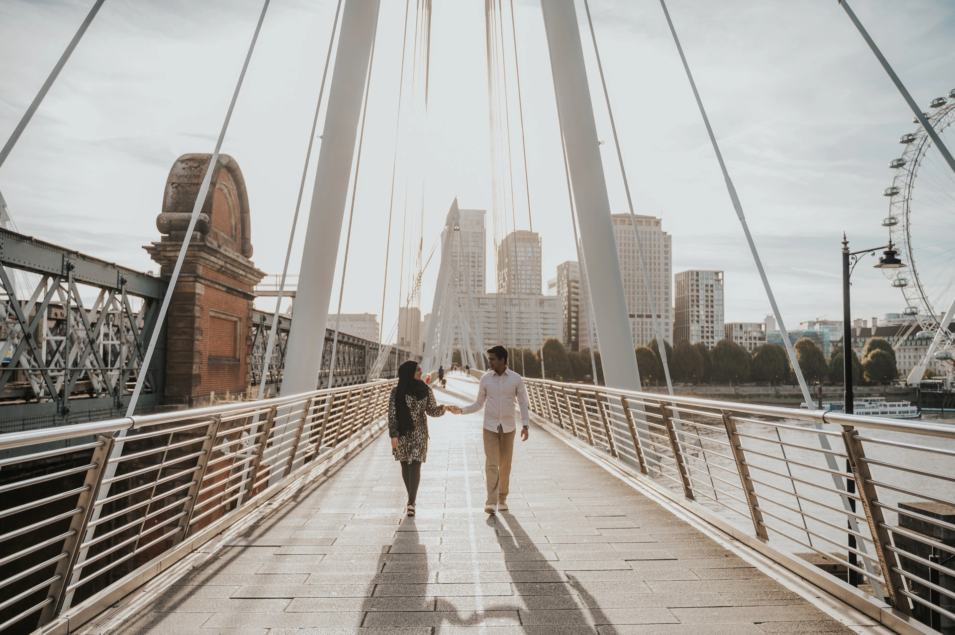 Walking together beneath the cables of the Golden Jubilee Bridge, with the London Eye at the edge of the view, during a London couple photoshoot.