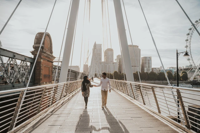 Walking together beneath the cables of the Golden Jubilee Bridge, with the London Eye at the edge of the view, during a London couple photoshoot.