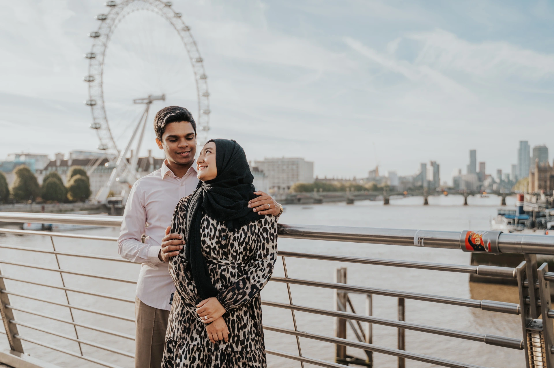 Wide portrait of the couple together by the Golden Jubilee Bridge railings, with the London Eye and Thames behind them, during their London photoshoot.