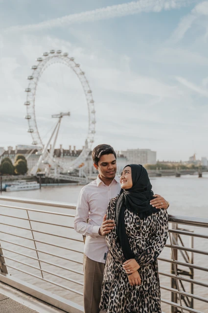 London couple photoshoot with the London Eye behind them Couple standing together on the Golden Jubilee Bridge, with her patterned dress and black headscarf beside his pale pink shirt, during their London photoshoot.
