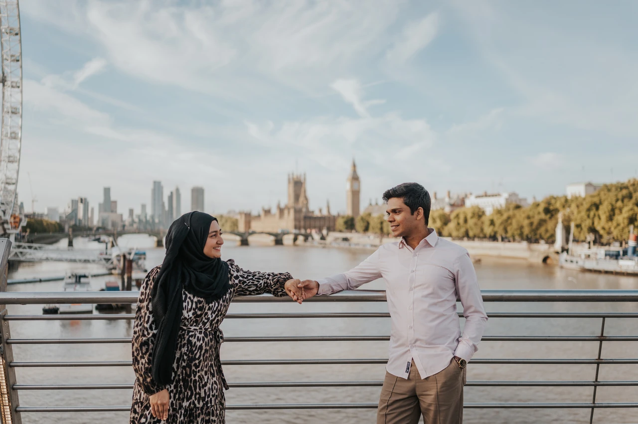 London couple photoshoot with Parliament across the river Couple holding hands beside the Golden Jubilee Bridge railings, with Parliament across the Thames, during their London photoshoot.