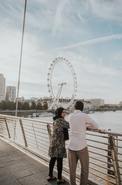 London couple photoshoot with a wide London Eye view Couple beside the Golden Jubilee Bridge railings, with the London Eye across the Thames, during their London photoshoot.