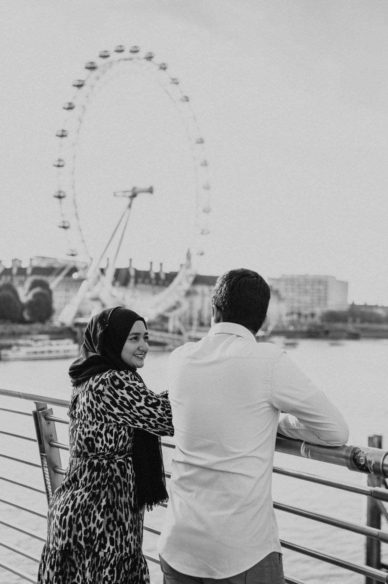 Black-and-white portrait of the couple at the Golden Jubilee Bridge railings, with the London Eye across the Thames, from their London photoshoot.