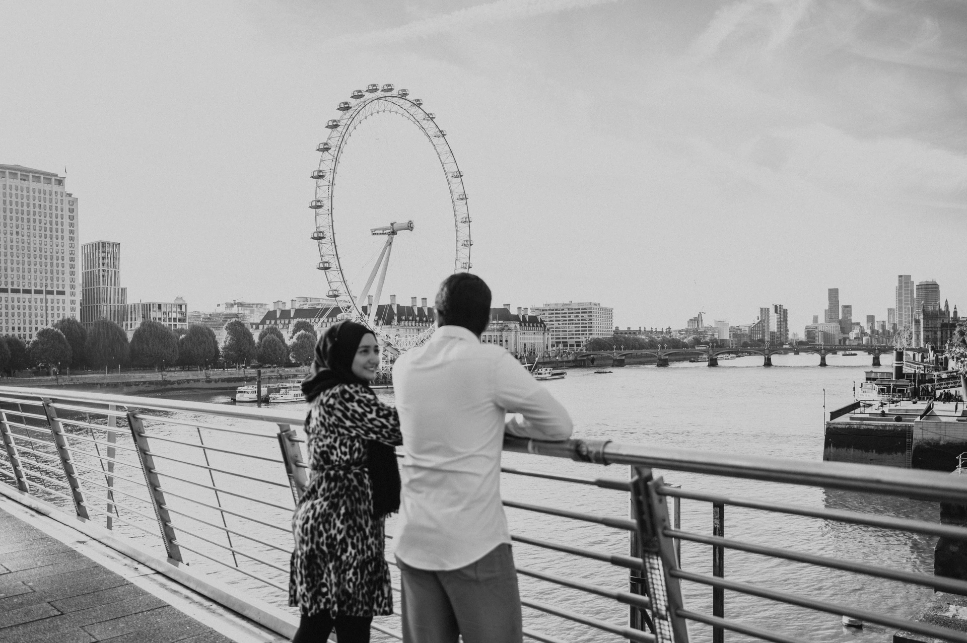 Wide black-and-white photograph of the couple beside the Golden Jubilee Bridge railings and the London Eye during their London photoshoot.