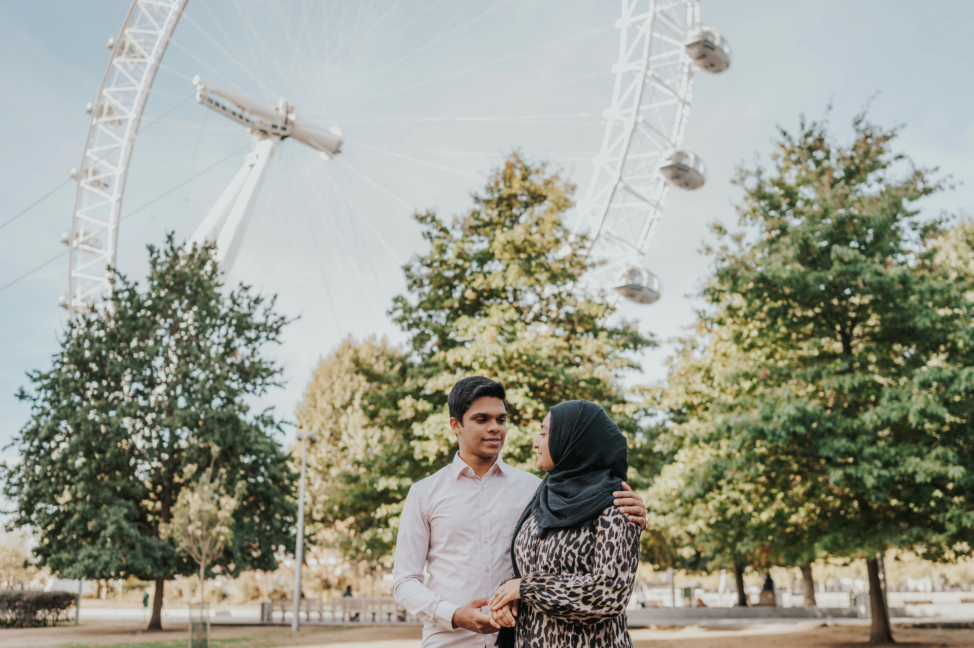 Couple standing close together beside the trees in Jubilee Gardens, beneath the London Eye, during their London photoshoot.