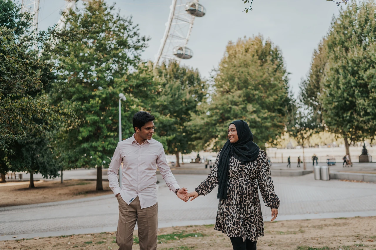 London couple photoshoot among the Jubilee Gardens trees Couple holding hands beside a curved path and trees in Jubilee Gardens, with London Eye capsules overhead, during their London photoshoot.