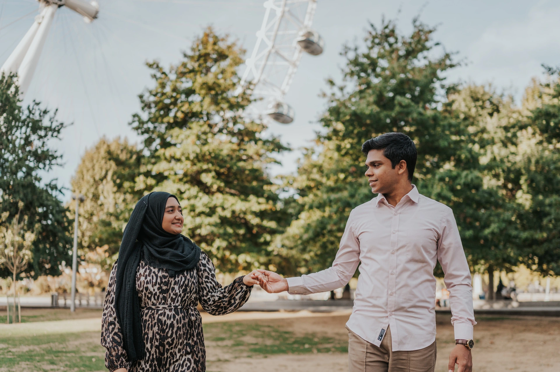 Couple holding hands in Jubilee Gardens, her patterned dress beside his pale pink shirt and the London Eye above the greenery, during their London photoshoot.