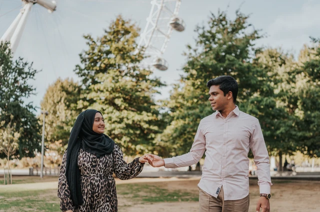 Couple holding hands in Jubilee Gardens, her patterned dress beside his pale pink shirt and the London Eye above the greenery, during their London photoshoot.