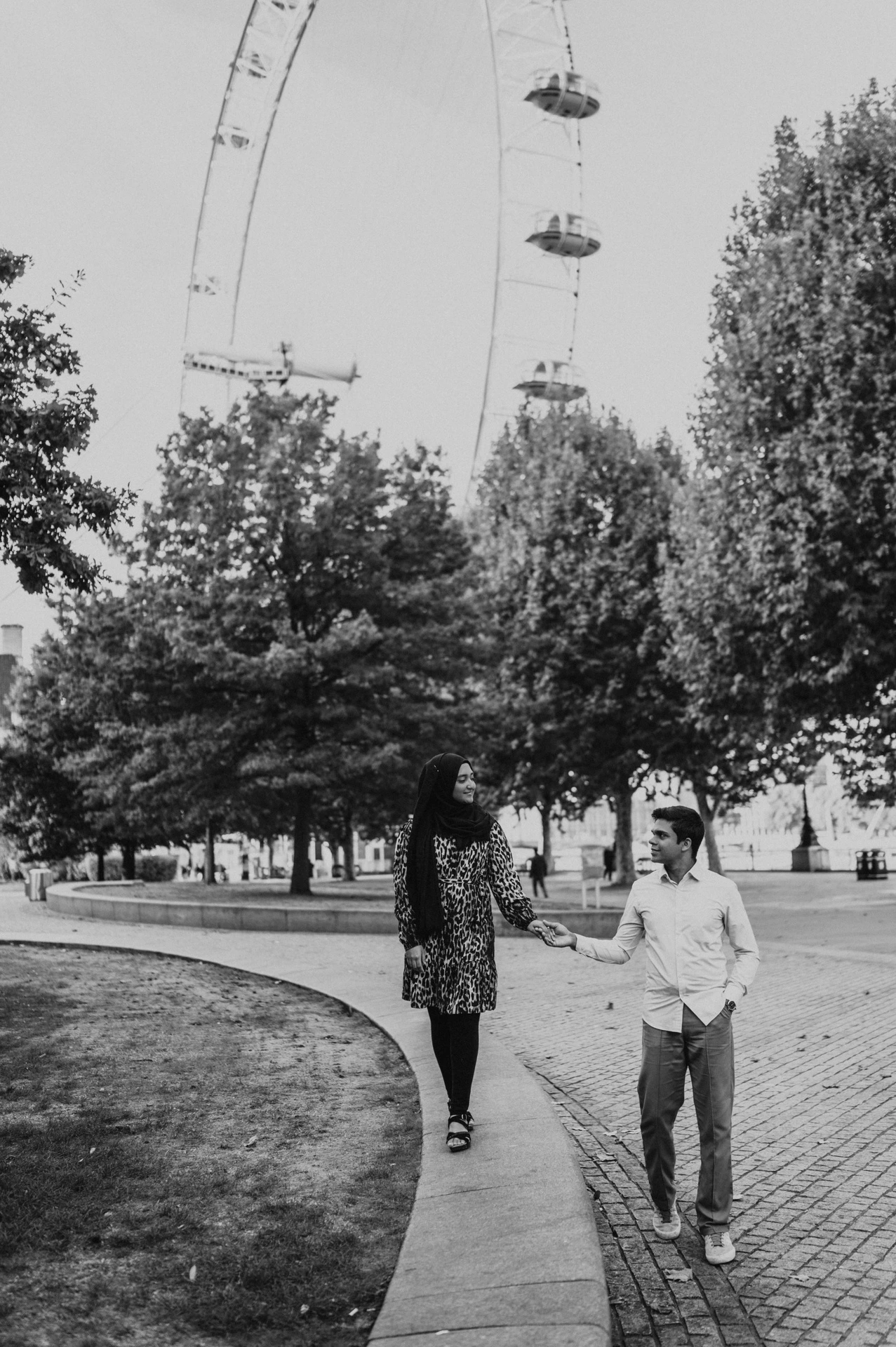 Black-and-white photograph of the couple holding hands on a Jubilee Gardens path beneath the London Eye during their London photoshoot.