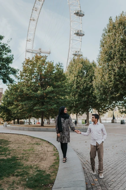 London couple photoshoot on the Jubilee Gardens path Full-length portrait of the couple holding hands on a path through Jubilee Gardens, with the London Eye above the trees, during their London photoshoot.